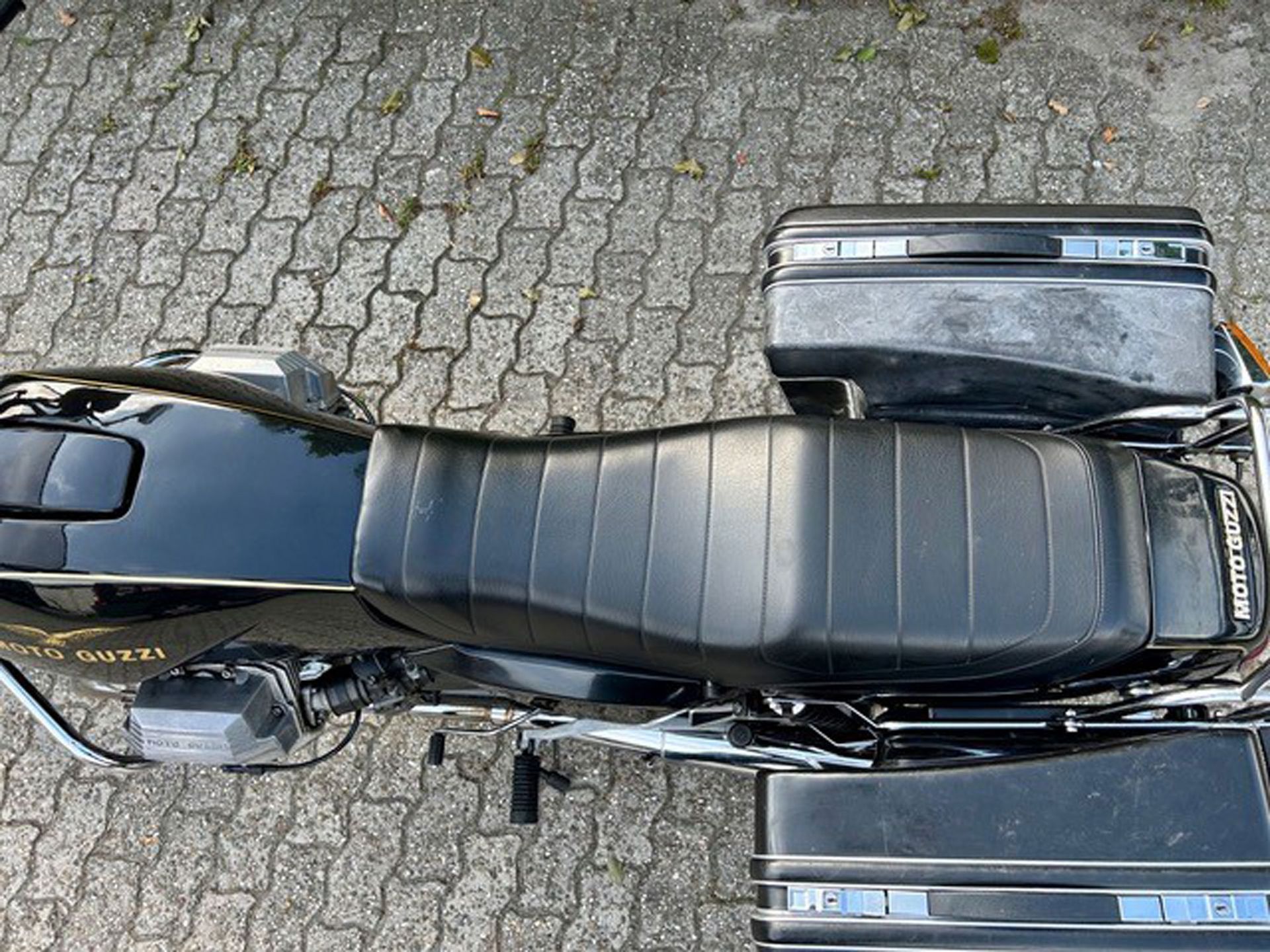 Schwarzes Moto Guzzi-Motorrad mit langem schwarzen Sitz und schwarzen Satteltaschen, geparkt auf einer gepflasterten Fläche.