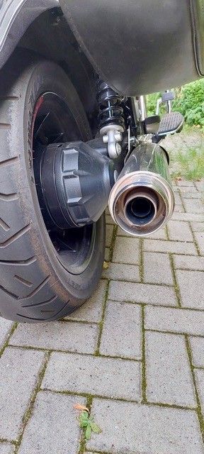 Nahaufnahme des Hinterreifens, des Auspuffs und der Federung eines Motorrads auf einem Ziegelweg.