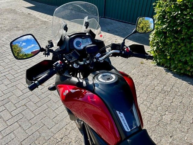 Ein rot-schwarzes Motorrad parkt auf einem sonnenbeschienenen Gehweg in der Nähe einer Hecke und eines Gebäudes.