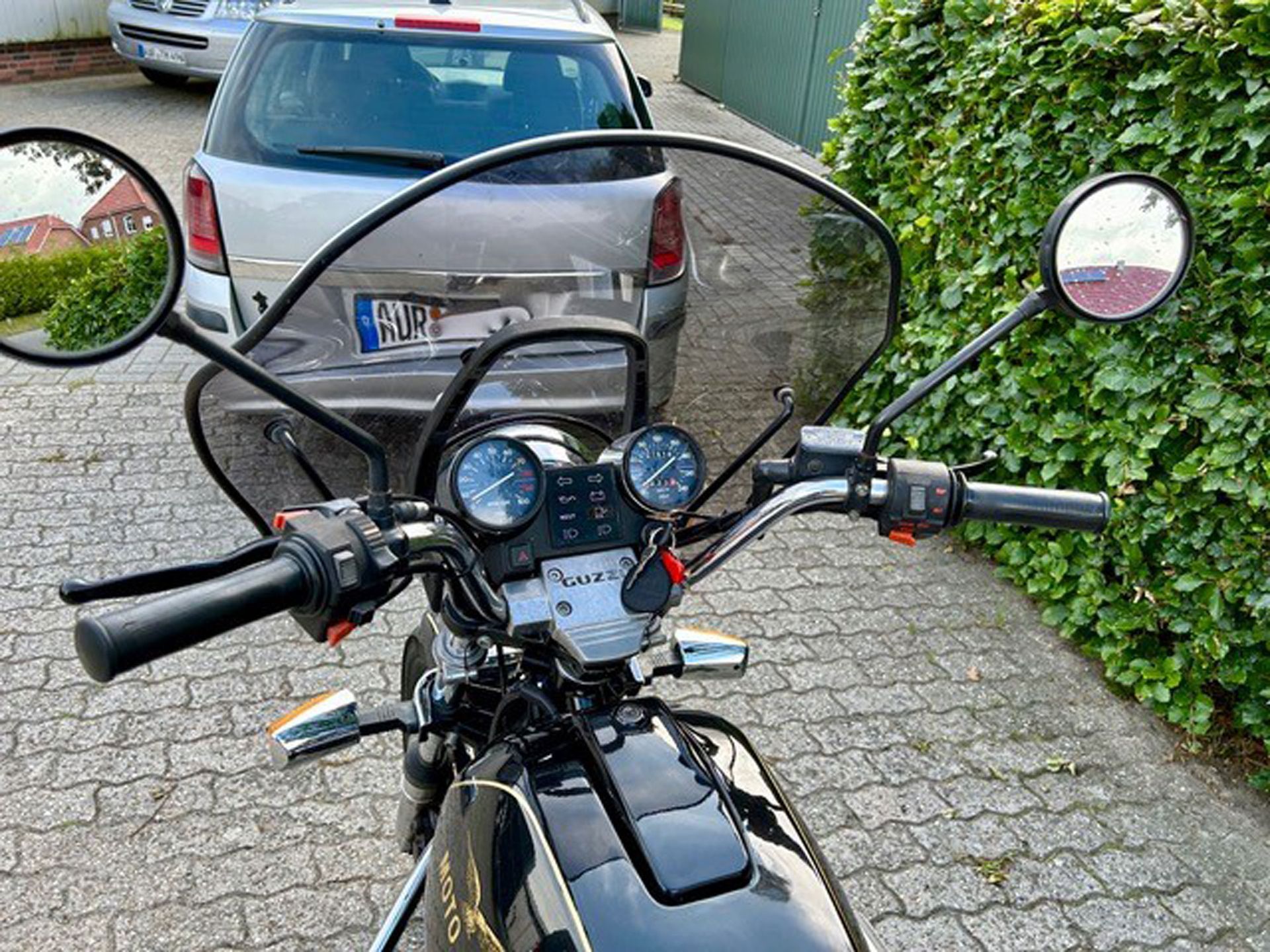 Blick vom Lenker eines schwarzen Motorrads mit Windschutzscheibe, Messgeräten, Spiegeln und einem Auto im Hintergrund.