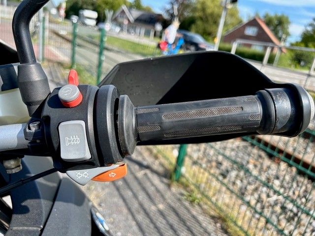 Motorradlenkergriff mit Bedienelementen, vor einem Zaun und Häusern.