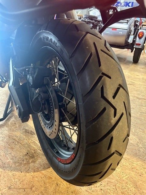 Nahaufnahme eines Motorradhinterreifens und einer Bremsscheibe in einem Ausstellungsraum, im Hintergrund ein weiteres Motorrad.