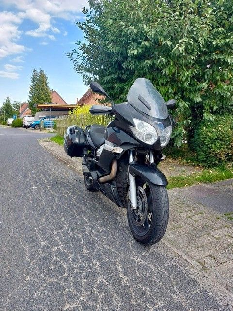 Schwarzes Motorrad mit Satteltaschen, geparkt auf einer asphaltierten Straße, Bäume und Häuser im Hintergrund.