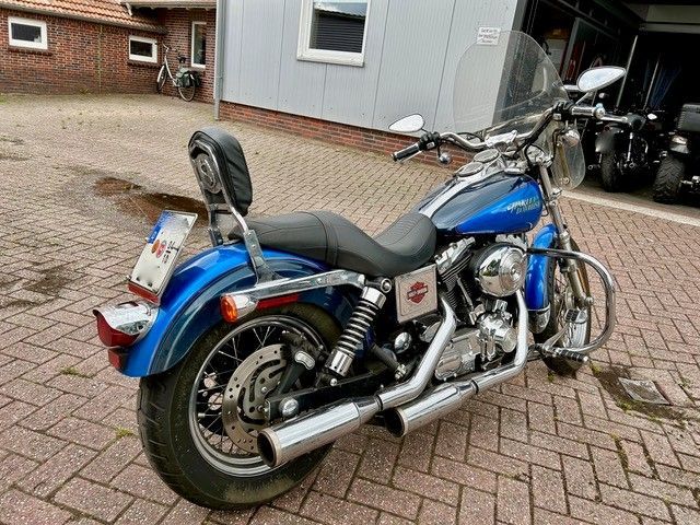 Blaues Harley-Davidson-Motorrad mit Rückenlehne, das auf einer Ziegelfläche vor einem Gebäude geparkt ist.