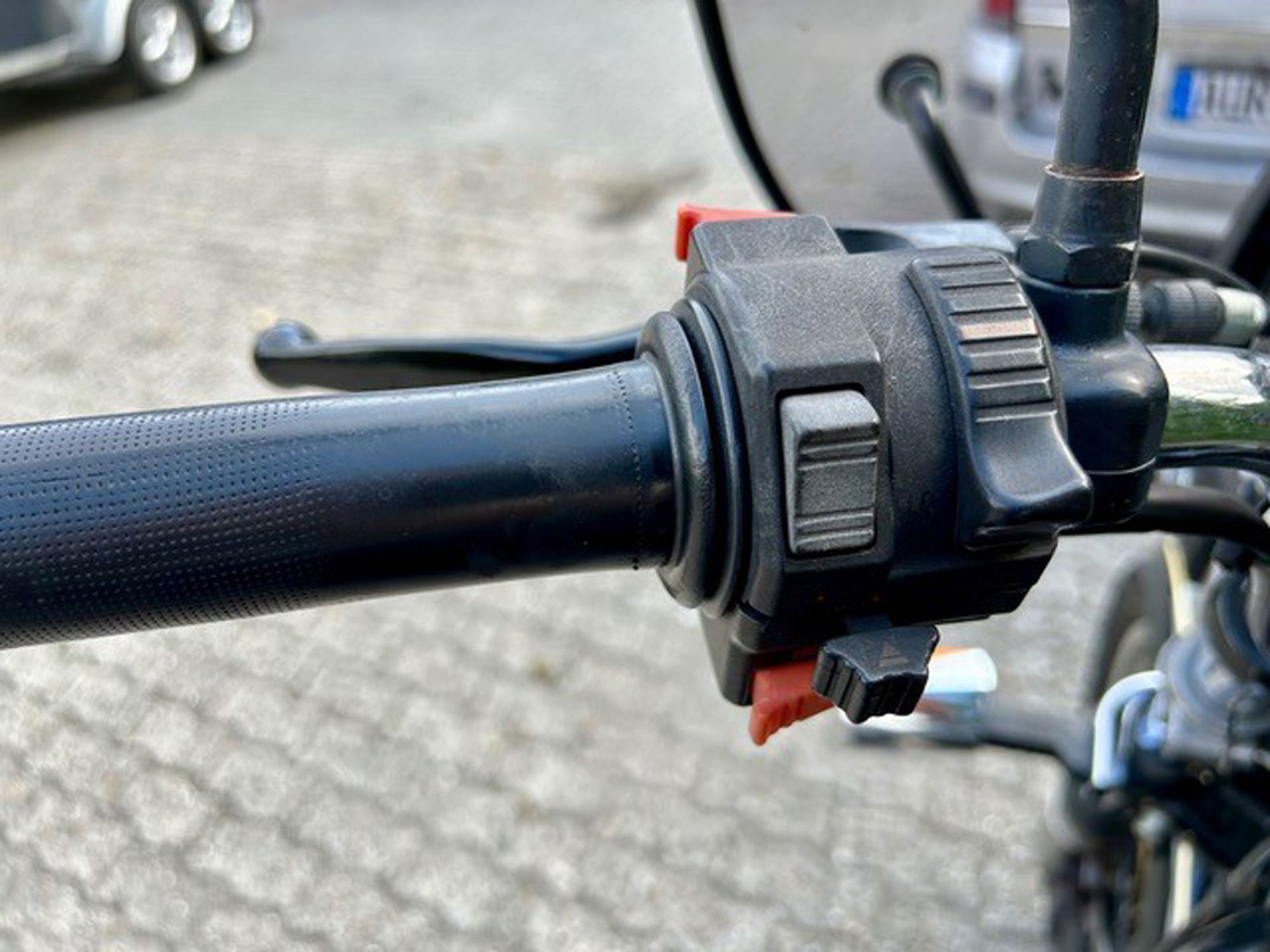 Schwarzer Motorradlenkergriff mit Bedienelementen einschließlich Schalter und roten Knöpfen.