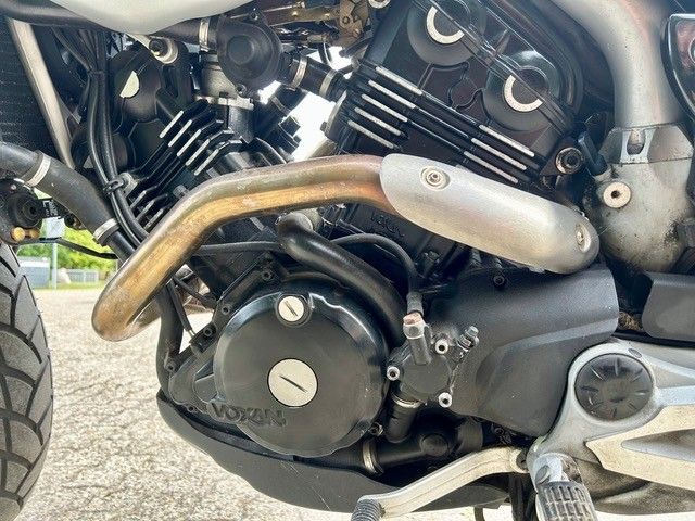 Nahaufnahme eines Motorradmotors mit schwarzem Gehäuse und gebogenem Auspuffrohr auf dem Asphalt.