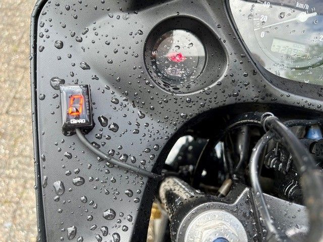 Schwarzes, regennass bespritztes Armaturenbrett mit einer kleinen Anzeige, einem orangefarbenen Schalter und freiliegenden Kabeln in der Nähe einer Radnabe.