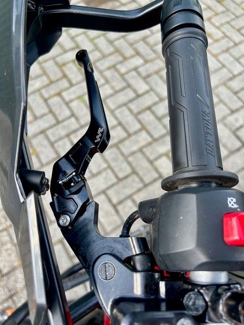 Nahaufnahme eines Fahrradlenkers mit schwarzem Bremshebel und rotem Schalter vor gepflastertem Hintergrund