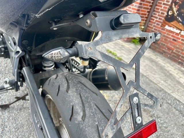 Nahaufnahme eines Motorradhinterreifens, Rücklichts und Kennzeichenhalters in der Nähe einer Backsteinmauer.