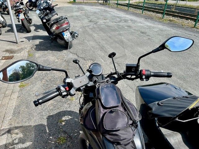 Motorradlenker mit Spiegeln und Tankrucksack, draußen mit anderen Motorrädern geparkt.