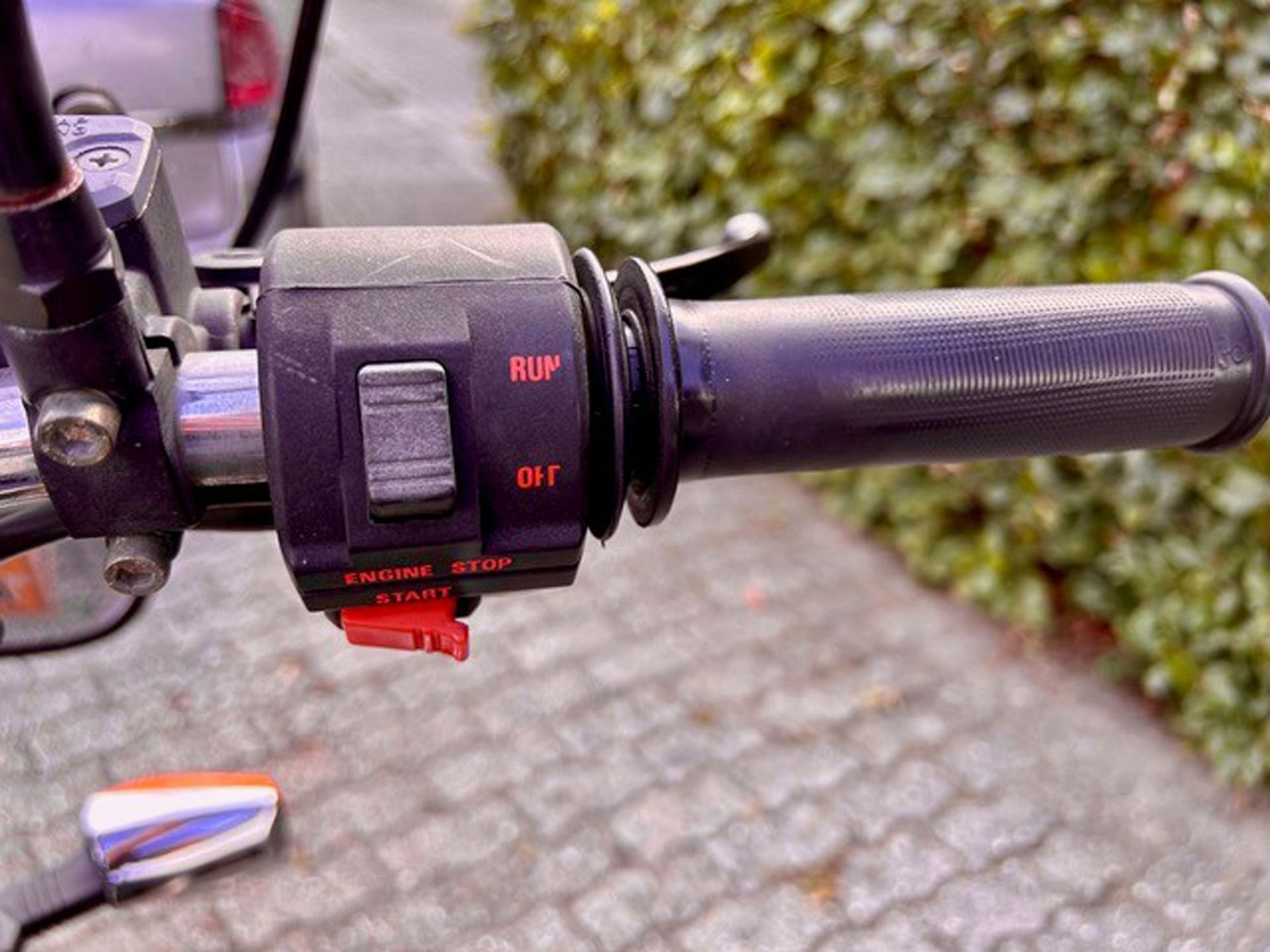 Nahaufnahme eines Motorradlenkers mit Bedienelementen, darunter ein Ein-/Ausschalter und ein roter Startknopf.
