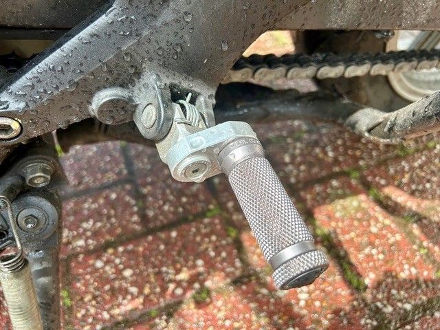Nahaufnahme einer Motorradfußraste mit einer silbernen, gerändelten Raste, die auf einem schwarzen Rahmen montiert ist.