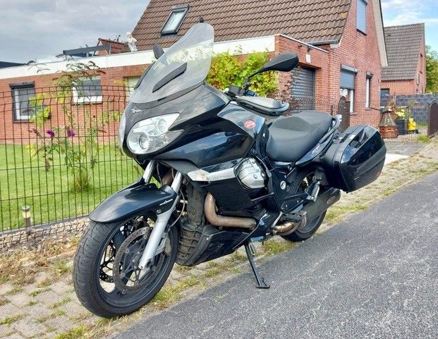Schwarzes Moto Guzzi-Tourenmotorrad, geparkt auf einer asphaltierten Straße neben einem Backsteinhaus mit Garten.