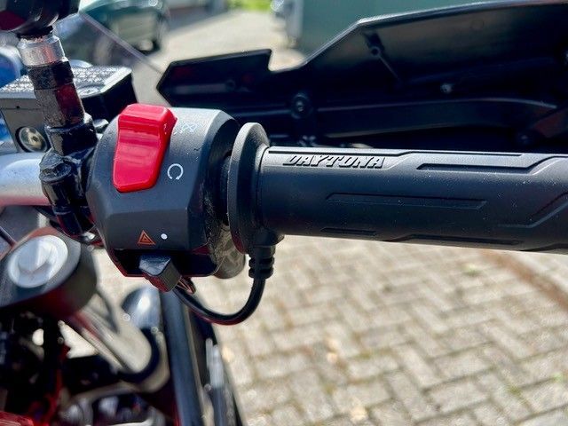 Nahaufnahme eines Motorradlenkers mit rotem Schalter und schwarzem Griff auf einer gepflasterten Straße