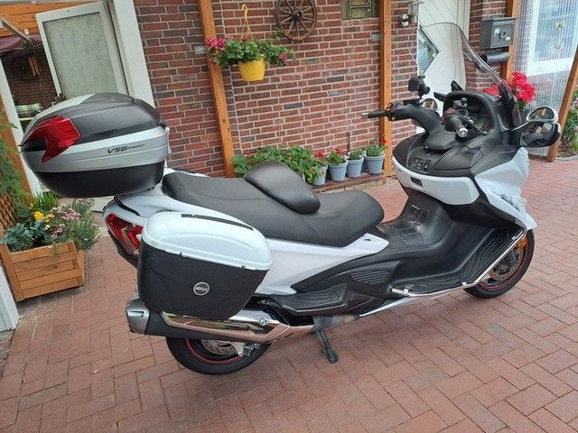 Weiß-schwarzer Motorroller mit seitlichen und oberen Ablagefächern, geparkt auf einer gemauerten Terrasse, mit einer Ziegelmauer und Blumen im Hintergrund.