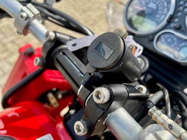 Nahaufnahme eines roten Motorradlenkers und Bedienelementen mit Instrumentenanzeigen im Hintergrund
