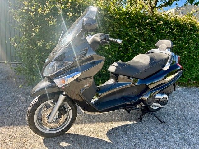 Ein blauer Maxi-Scooter parkt auf einer sonnigen Einfahrt neben einer Hecke.