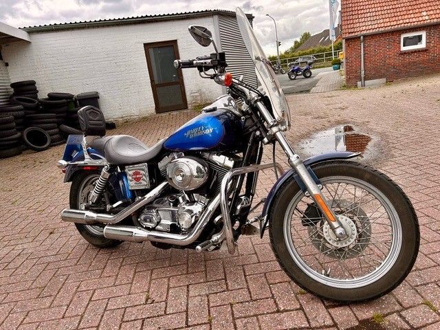 Blaues Harley-Davidson-Motorrad auf Ziegelpflaster geparkt. Silberner Motor, verchromter Auspuff und Windschutzscheibe vor einem Gebäude aus Ziegeln und weißen Wänden.