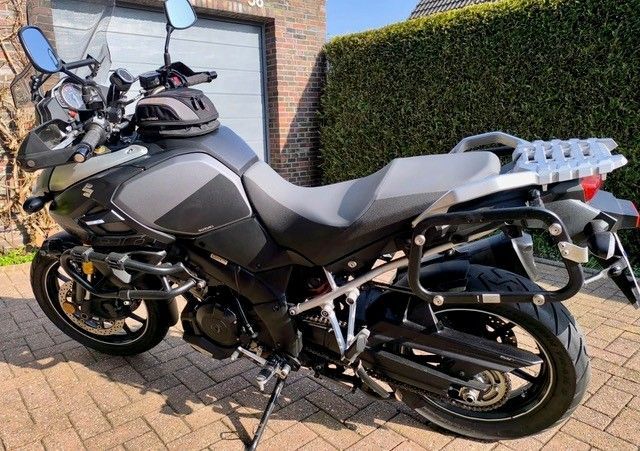 Graues Suzuki V-Strom-Motorrad, das an einem sonnigen Tag auf einer gemauerten Auffahrt vor einer Garage und Büschen geparkt ist.
