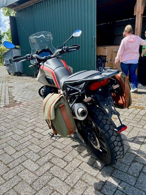 Ein Motorrad mit Packtaschen steht auf einer gepflasterten Auffahrt neben einem blauen Schuppen; eine Frau geht in der Nähe entlang.