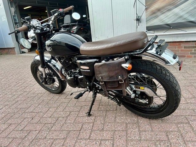 Schwarzes Motorrad mit braunem Sitz, geparkt auf einem gepflasterten Gehweg neben einem Gebäude