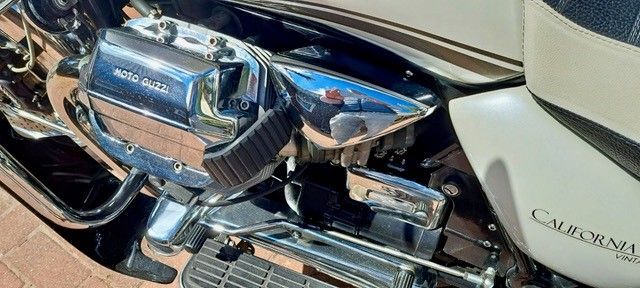Nahaufnahme eines glänzenden Motorradmotors aus Chrom mit dem sichtbaren Wort „Moto Guzzi“ und einer weißen Seitenverkleidung mit der Aufschrift „California“.