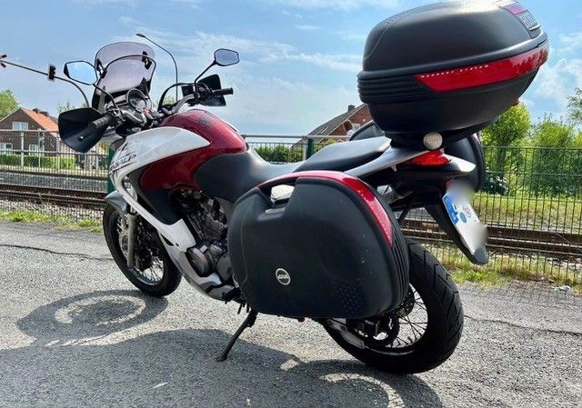 Ein rot-weißes Motorrad mit Satteltaschen und Topcase, das neben Bahngleisen geparkt ist.