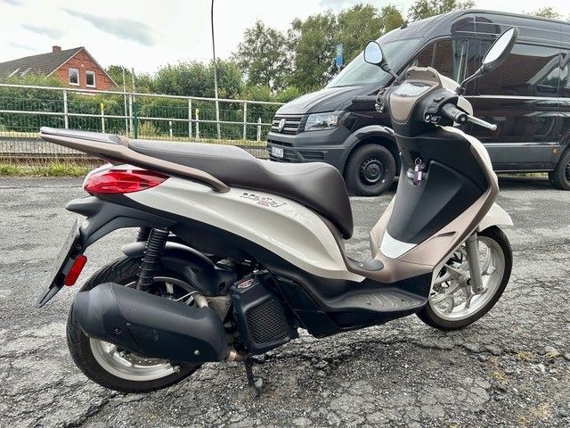 Ein silberner Roller steht auf einem rissigen Parkplatz, im Hintergrund sind ein schwarzer Lieferwagen und eine Leitplanke zu sehen.