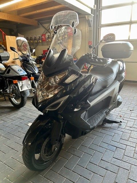 Ein schwarzer Roller steht in einer Garage; dahinter befindet sich eine durchsichtige Windschutzscheibe und ein Topcase.