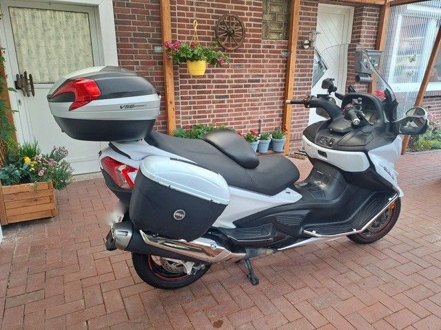 Ein weiß-schwarzer Roller mit Staufächern steht auf einer gemauerten Terrasse. Der Roller steht vor einer roten Ziegelmauer und -tür.