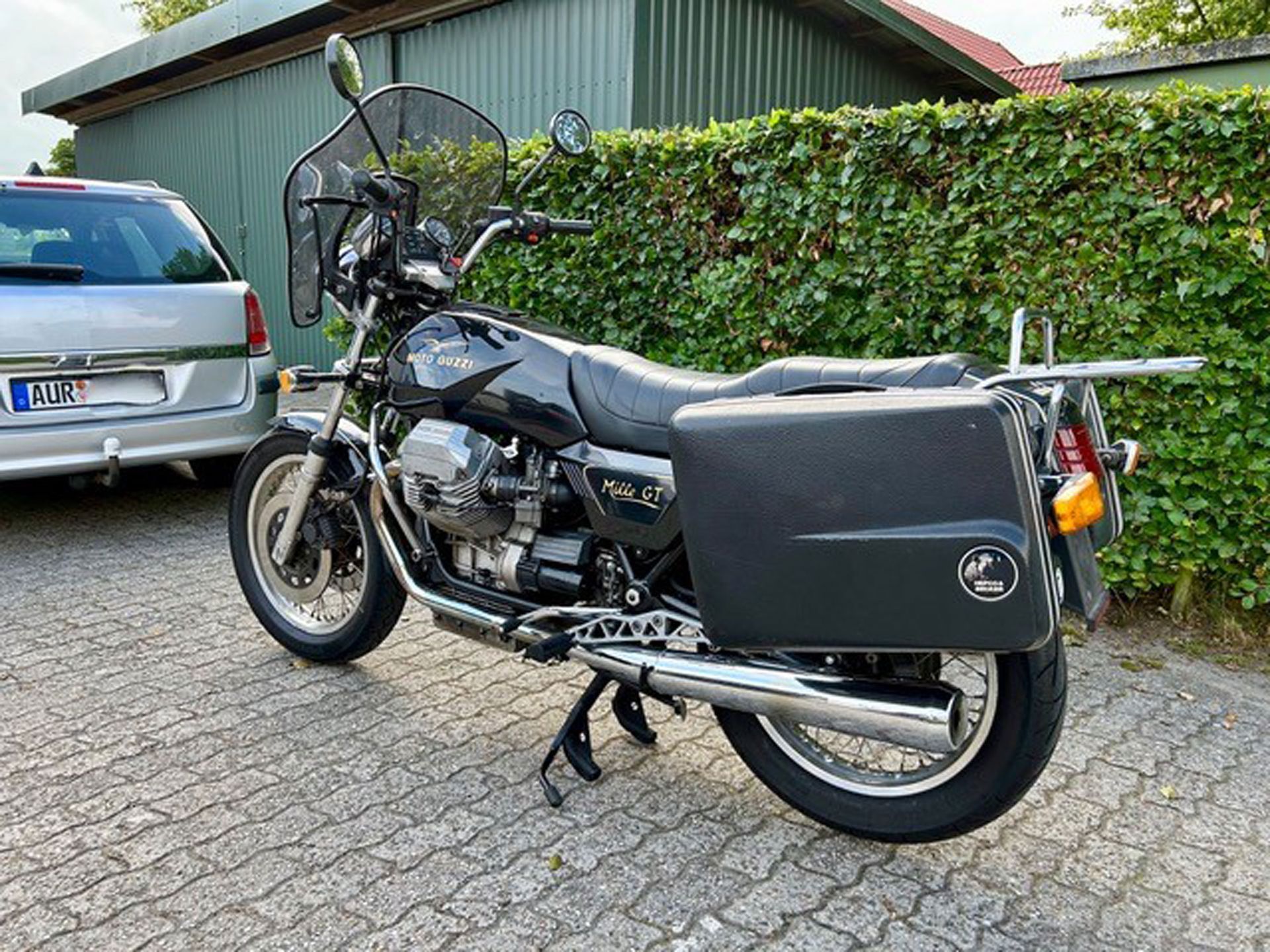 Schwarzes Moto Guzzi-Motorrad mit Seitenkoffern, geparkt auf einer gepflasterten Fläche, im Hintergrund ein silbernes Auto und eine grüne Hecke.