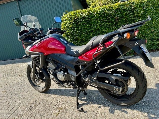 Ein rot-schwarzes Motorrad steht auf dem Bürgersteig neben einer grünen Hecke und einem blauen Gebäude, Seitenansicht.