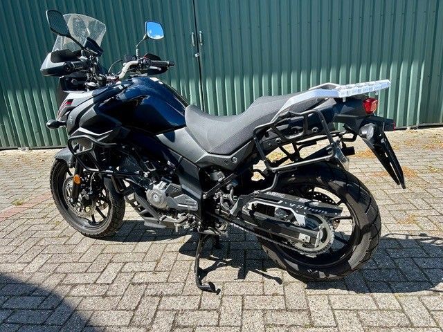 Ein blaues Tourenmotorrad parkt auf einer gepflasterten Auffahrt neben einer grünen Wellblechmauer.