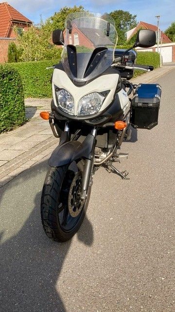 Schwarz-weißes Suzuki V-Strom-Motorrad, geparkt auf einer asphaltierten Straße; schwarzer Seitenkoffer angebracht, mit Grün und Häusern im Hintergrund.