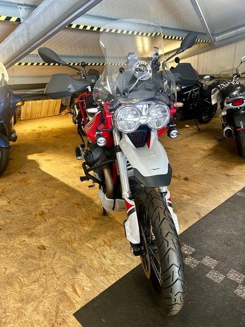 Motorrad mit grauer Karosserie und roten Akzenten, das in einer Halle zwischen anderen Motorrädern geparkt ist, Vorderansicht.