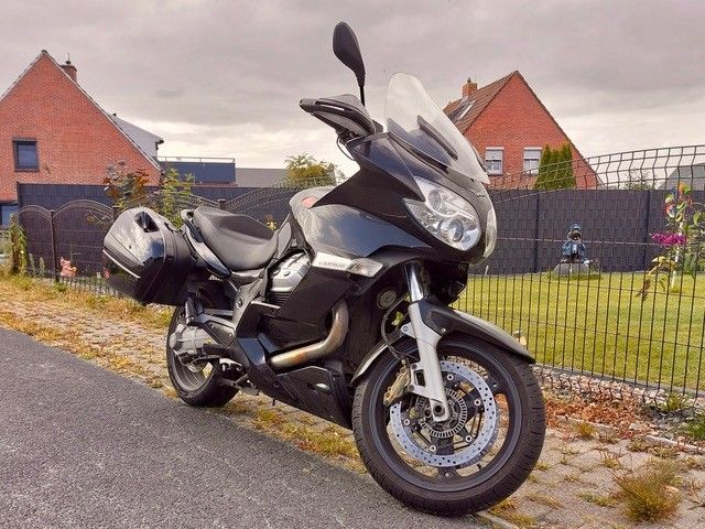 Schwarzes Moto Guzzi-Motorrad, geparkt auf einem Weg vor einem Zaun und Backsteinhäusern.