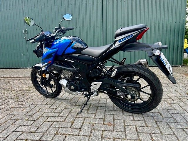 Ein blau-schwarzes Sportmotorrad parkt auf einem Backsteinpflaster neben einer grünen Wellblechwand.