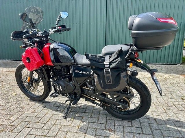 Schwarzes Adventure-Motorrad mit roter Frontabdeckung und Topcase, geparkt auf einem gepflasterten Parkplatz vor einer grünen Mauer.