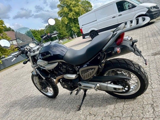 Schwarzes Motorrad parkt auf einer Straße, im Hintergrund sind ein weißer Lieferwagen und Bäume zu sehen.