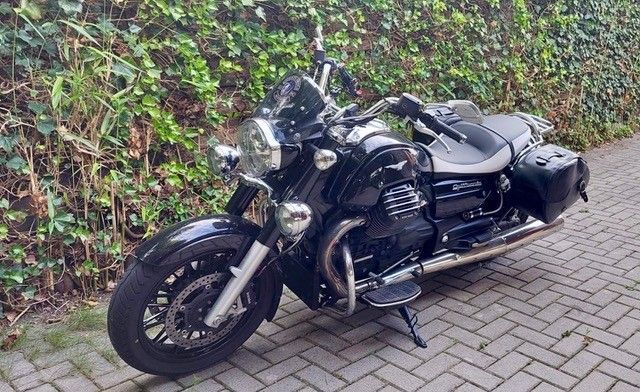 Schwarzes Moto Guzzi-Motorrad auf Ziegelstein geparkt, mit einer grünen Hecke im Hintergrund.