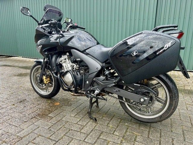 Schwarzes Tourenmotorrad parkt auf einer gepflasterten Fläche neben einer grünen Wellblechwand.