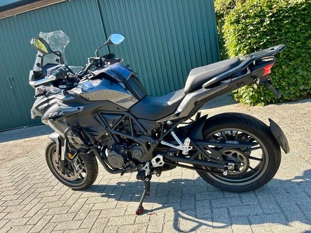 Ein blaues Motorrad parkt auf einer gepflasterten Auffahrt neben einem grünen Zaun und einer Hecke.