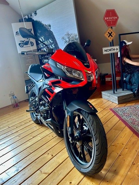 Ein rot-schwarzes Sportmotorrad steht in einem Innenraum auf einem Holzboden, neben einem alten Verkehrsschild und einem roten Teppich.