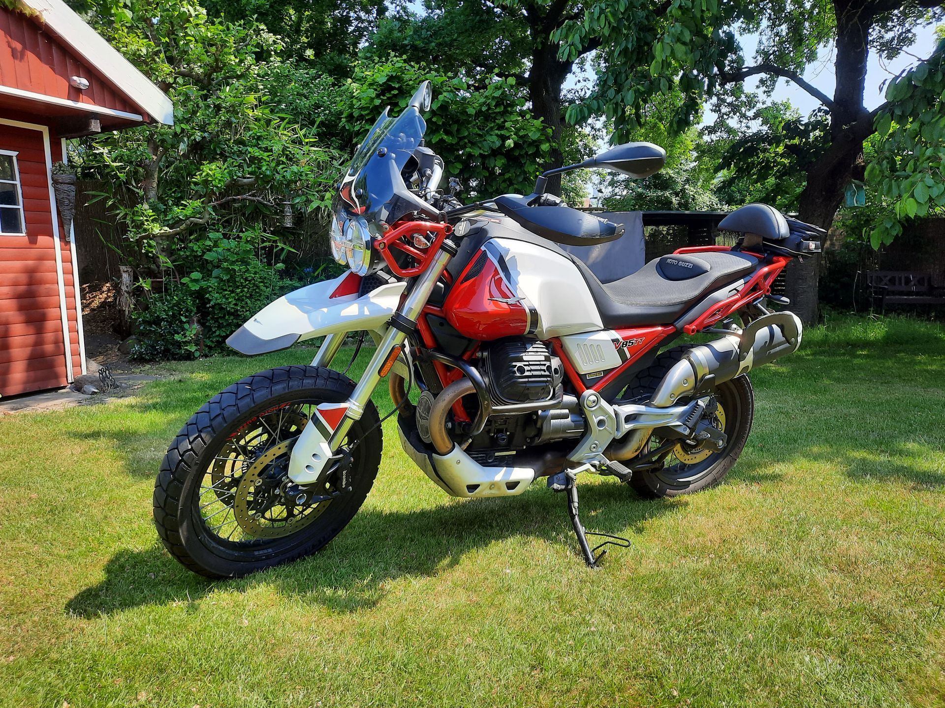 Ein rot-weißes Motorrad steht im Gras vor einem roten Schuppen.