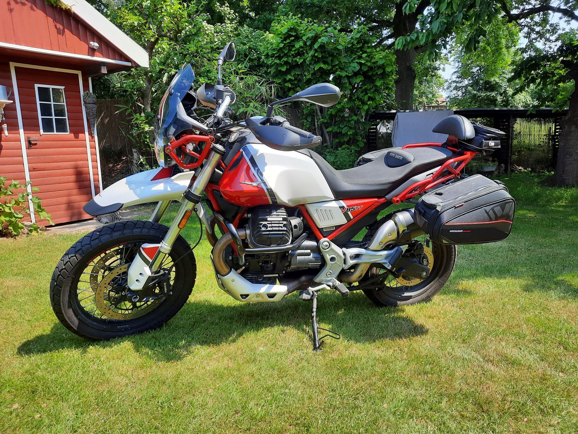 Ein rot-weißes Motorrad steht im Gras vor einem roten Schuppen.