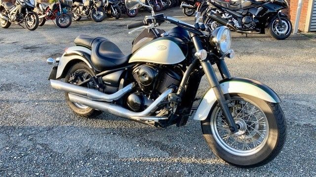 Ein silber-schwarzes Kawasaki Vulcan Motorrad parkt auf Asphalt, im Hintergrund sind weitere Motorräder zu sehen.