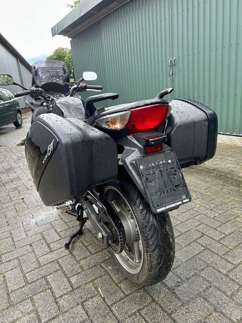 Schwarzes Tourenmotorrad parkt auf einer nassen Ziegelsteinauffahrt neben einem grünen Gebäude.