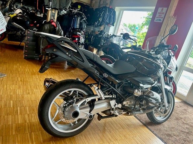 Schwarzes Sportmotorrad parkt in einem Innenraum auf einem Holzboden, im Hintergrund sind weitere Motorräder zu sehen.