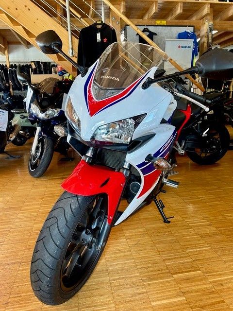 Ein rot-weiß-blaues Sportmotorrad ist in einem Ausstellungsraum mit anderen Motorrädern dahinter ausgestellt.