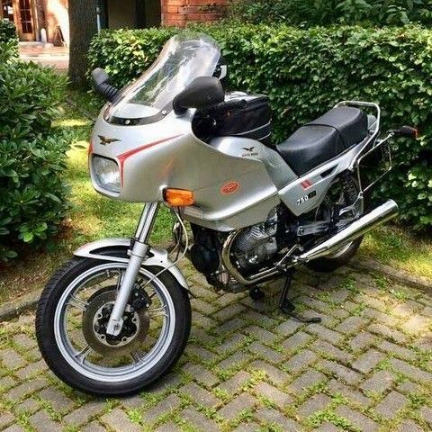 Ein silbernes Tourenmotorrad parkt auf einem Kopfsteinpflasterweg neben üppigem Grün.